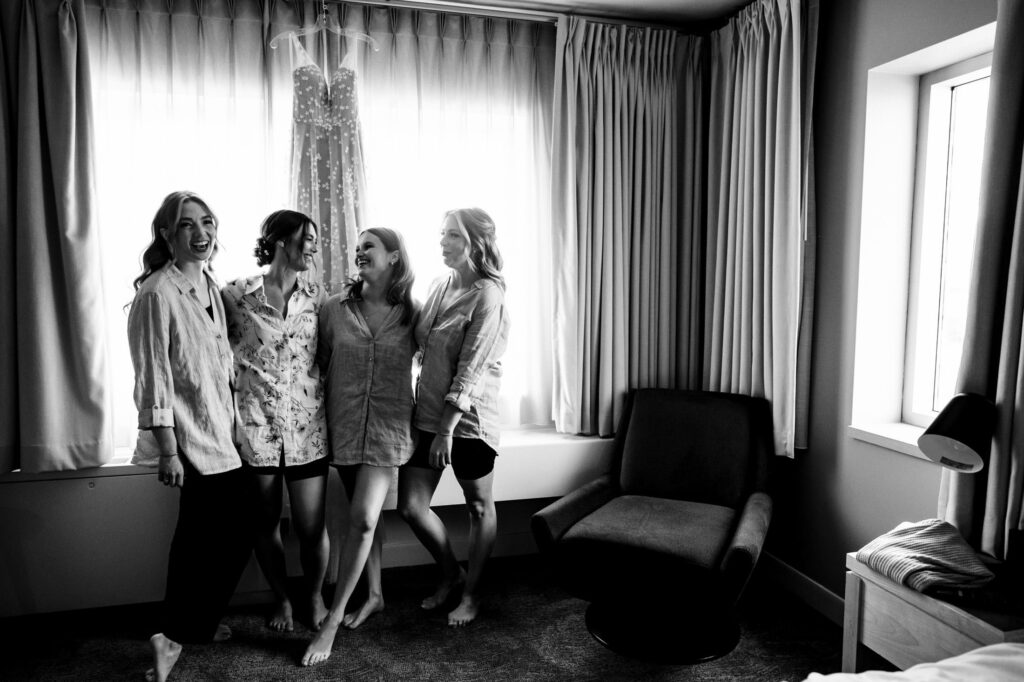 Four women smiling and laughing by a window with a dress hanging in the background.