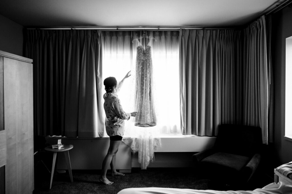 Person reaching towards a dress hanging in front of a window, in a dimly lit room.
