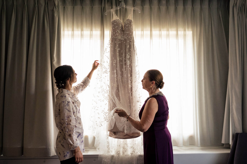 Two women admire a wedding dress hanging in front of a softly lit window.