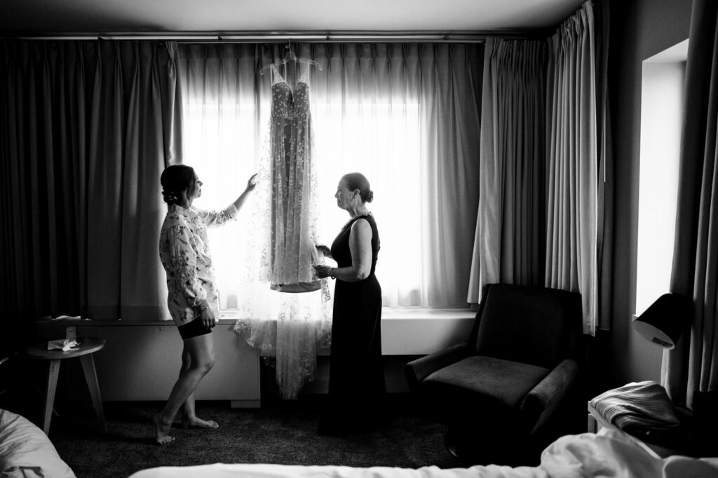 Two women admire a dress hanging by a window in a softly lit room.
