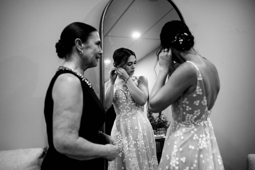 Bride adjusts earring in mirror as woman in black dress looks on.