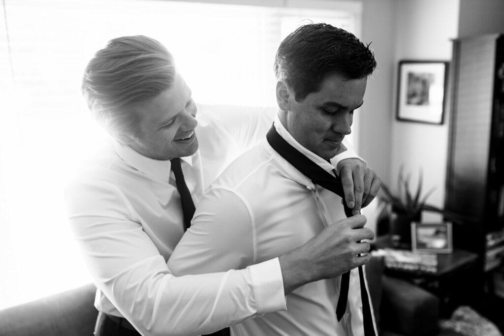 Two men in shirts and ties, one helping the other adjust his tie, smiling in a bright room.