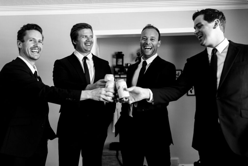 Four men in suits clinking beer cans, smiling and laughing indoors.