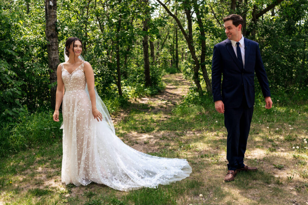 A bride and groom stand together on a forest path, surrounded by green trees.