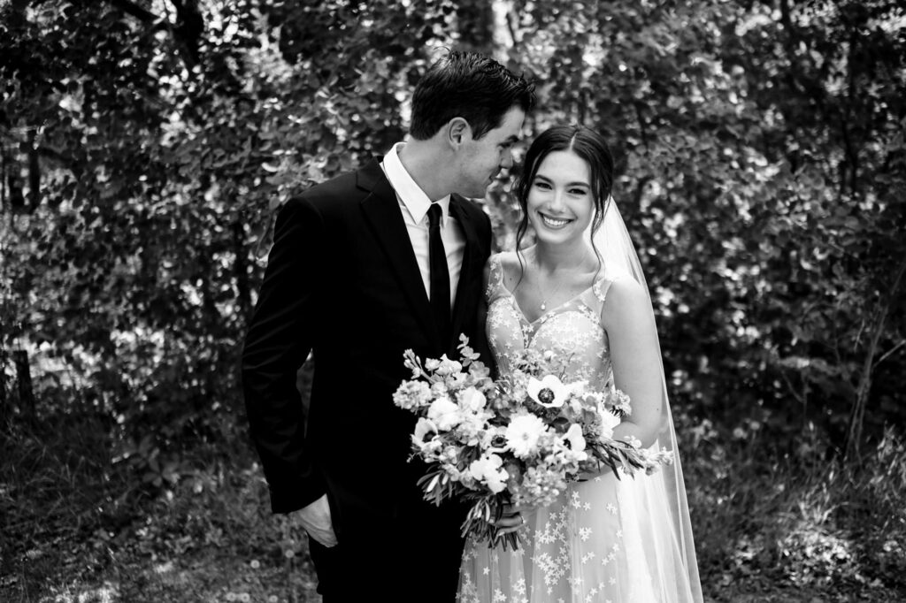 Bride holding bouquet, groom in suit, smiling in wooded area. Black and white photo.