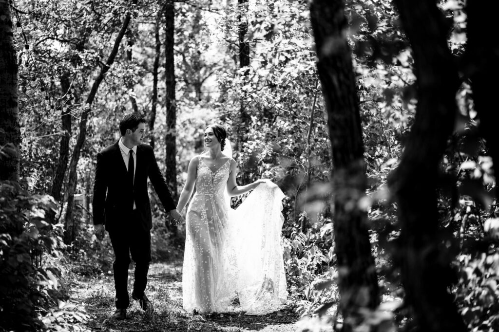 Couple in wedding attire walks through a sunlit forest path holding hands, joyful and smiling.