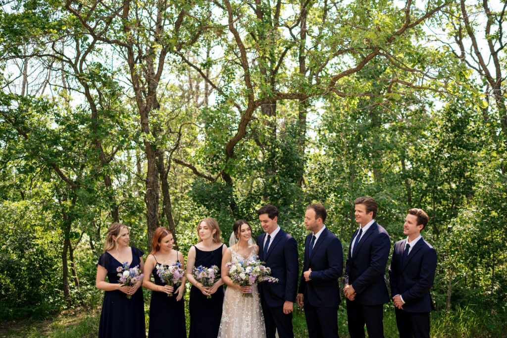 Wedding party in a forest, with the bride and groom surrounded by attendants in dark outfits.