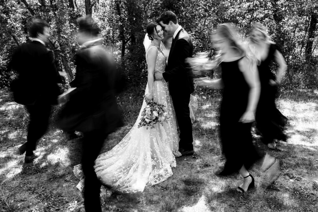 Bride and groom kiss in the center, blurred figures move around them, outdoor setting.