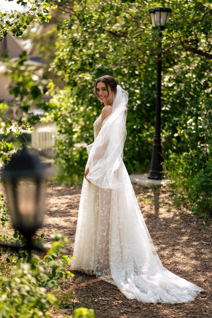 Bride in a white dress and veil stands outdoors, surrounded by lush greenery.