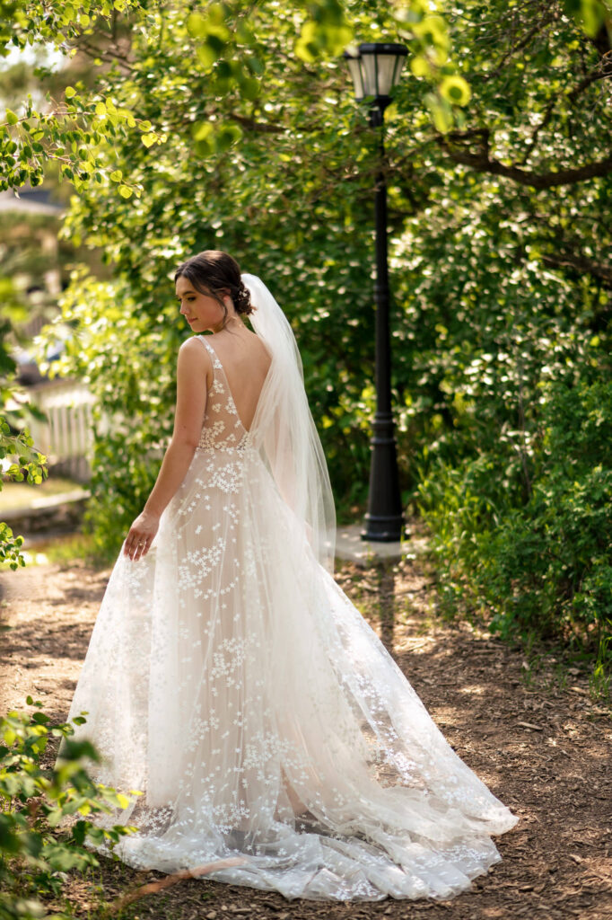 Bride in lace gown and veil stands on a wooded path, illuminated by sunlight.