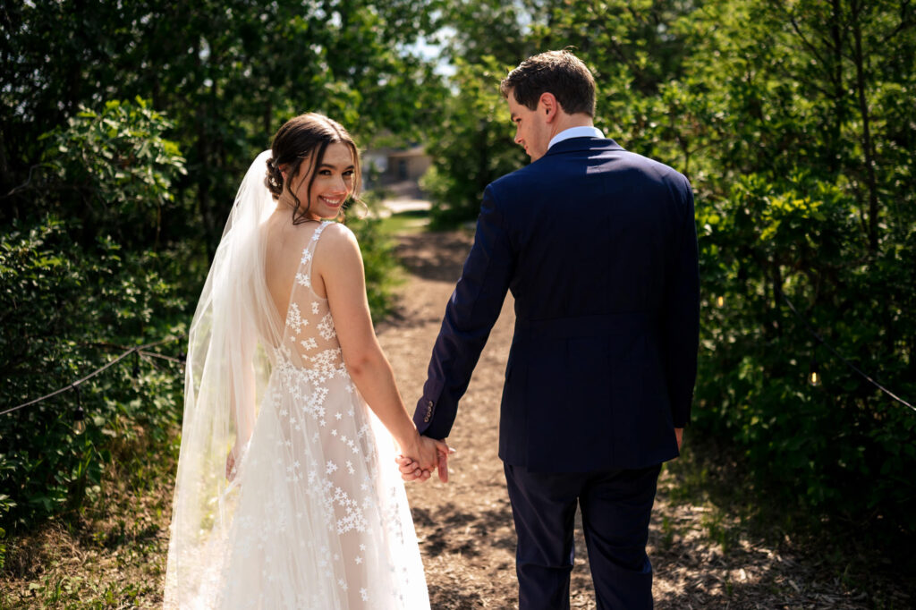 Bride and groom holding hands on a sunlit path, surrounded by greenery.