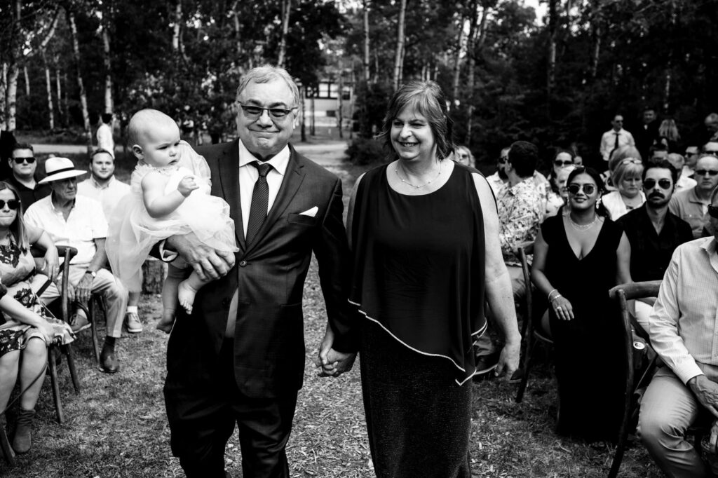 A couple walks down an outdoor aisle holding a baby, with guests seated around.