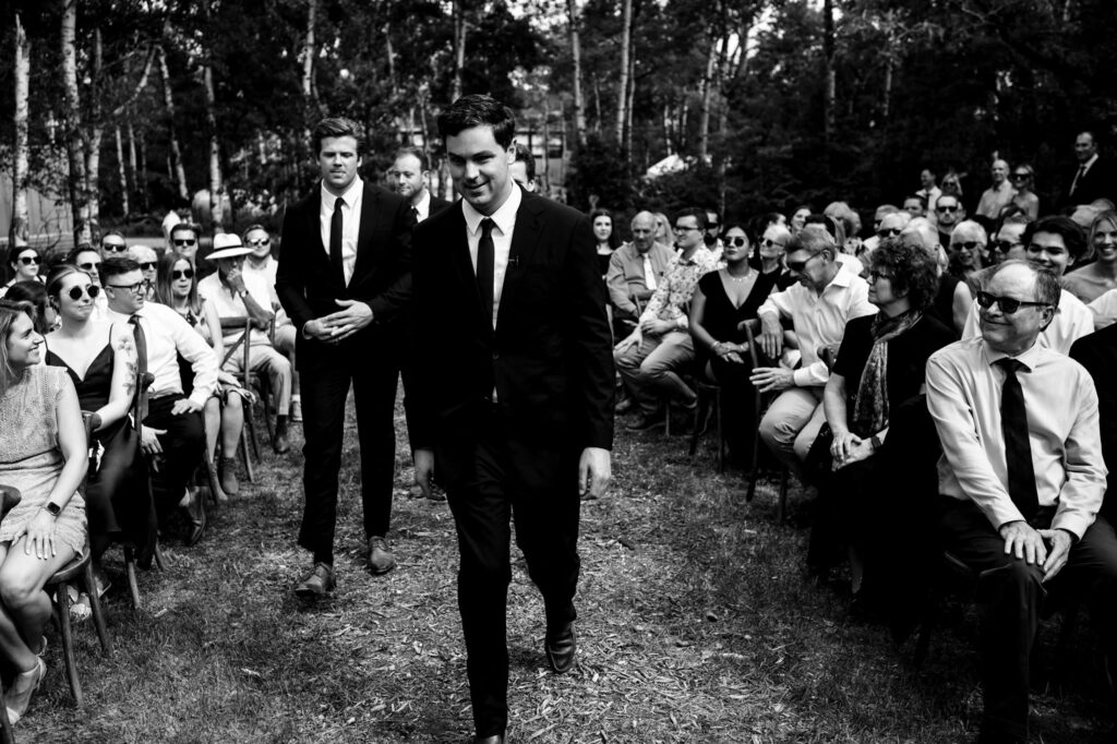A person in a suit walks down an aisle outdoors, surrounded by seated guests.