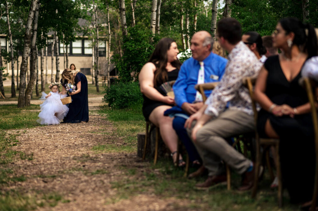 Children walking down an outdoor aisle at a wedding, guests seated on wooden chairs.