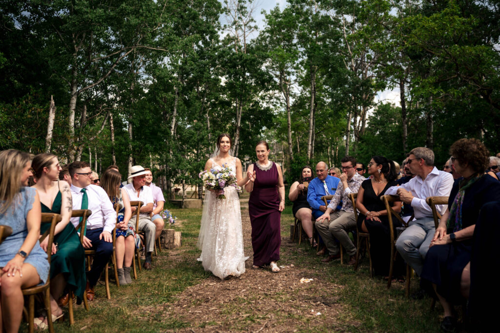 Bride walking down an outdoor aisle with a companion, surrounded by seated guests.