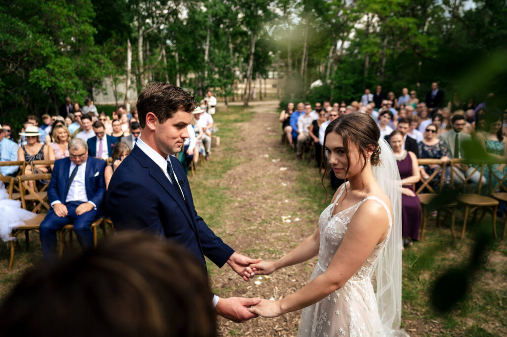 A couple holding hands during an outdoor wedding ceremony, with guests seated behind them.