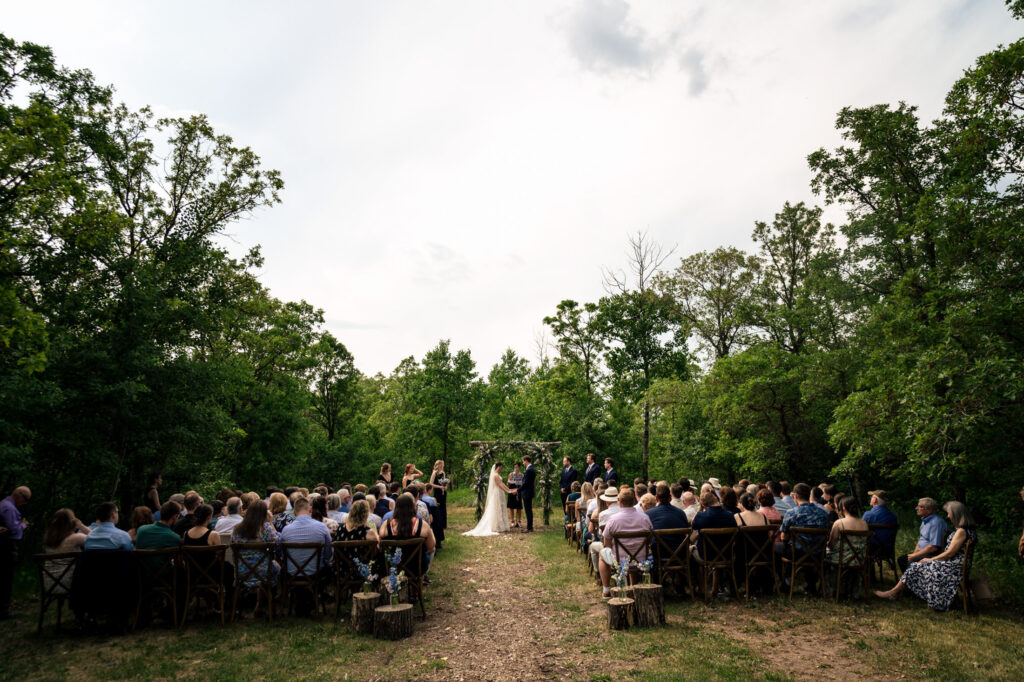 Outdoor wedding ceremony in a forest with guests seated on both sides of the aisle.