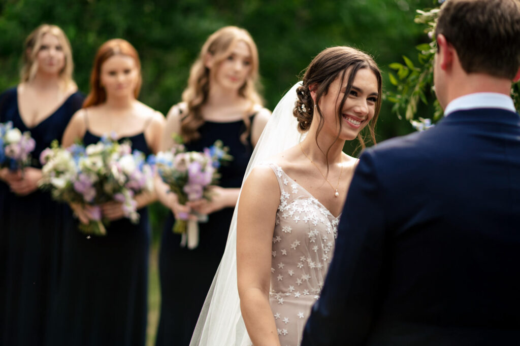 Bride smiling at groom during outdoor wedding ceremony, with bridesmaids holding bouquets in background.