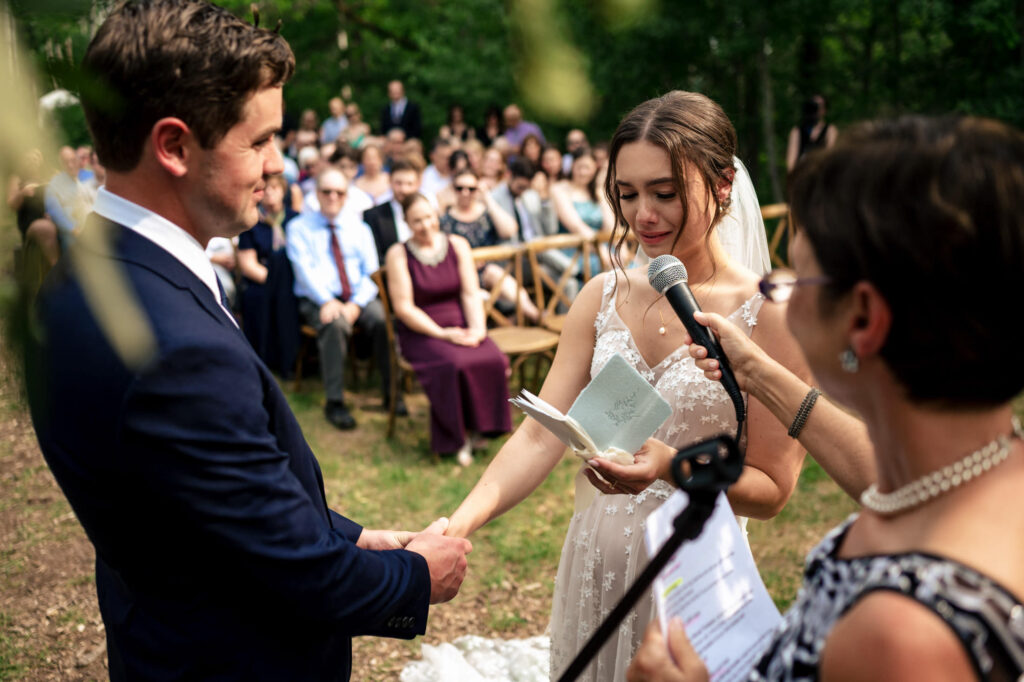 Bride reads vows to groom during an outdoor wedding ceremony, with guests seated in the background.