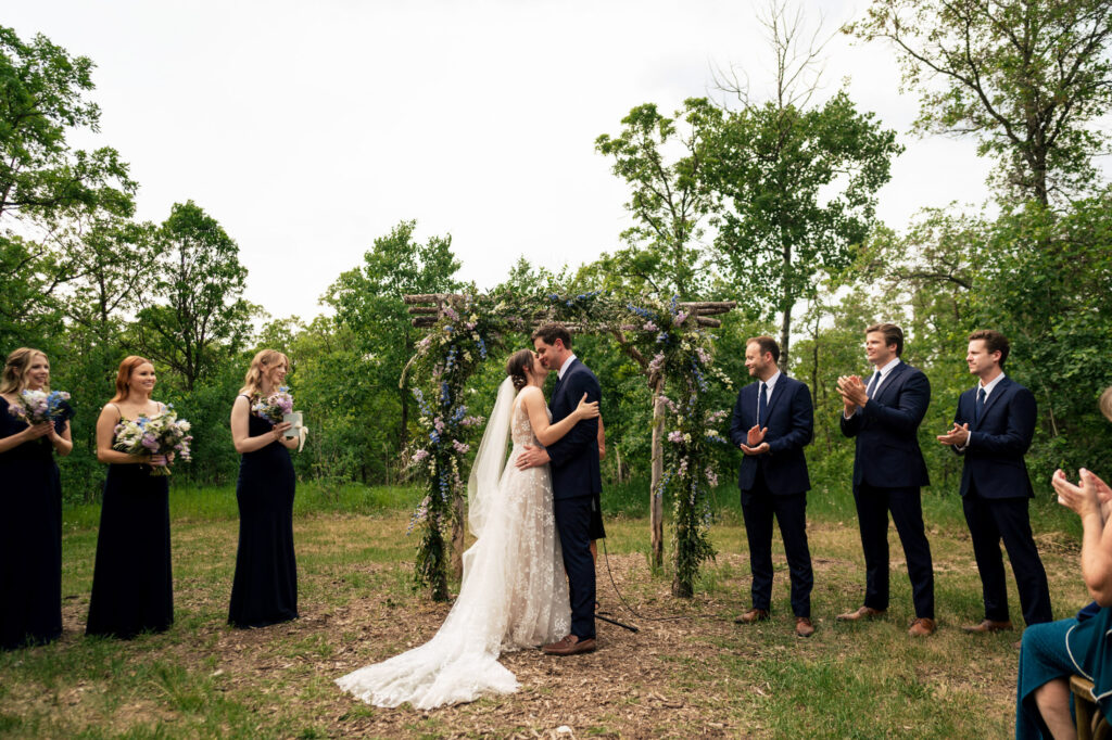 Bride and groom kiss at outdoor wedding ceremony, surrounded by bridesmaids and groomsmen.