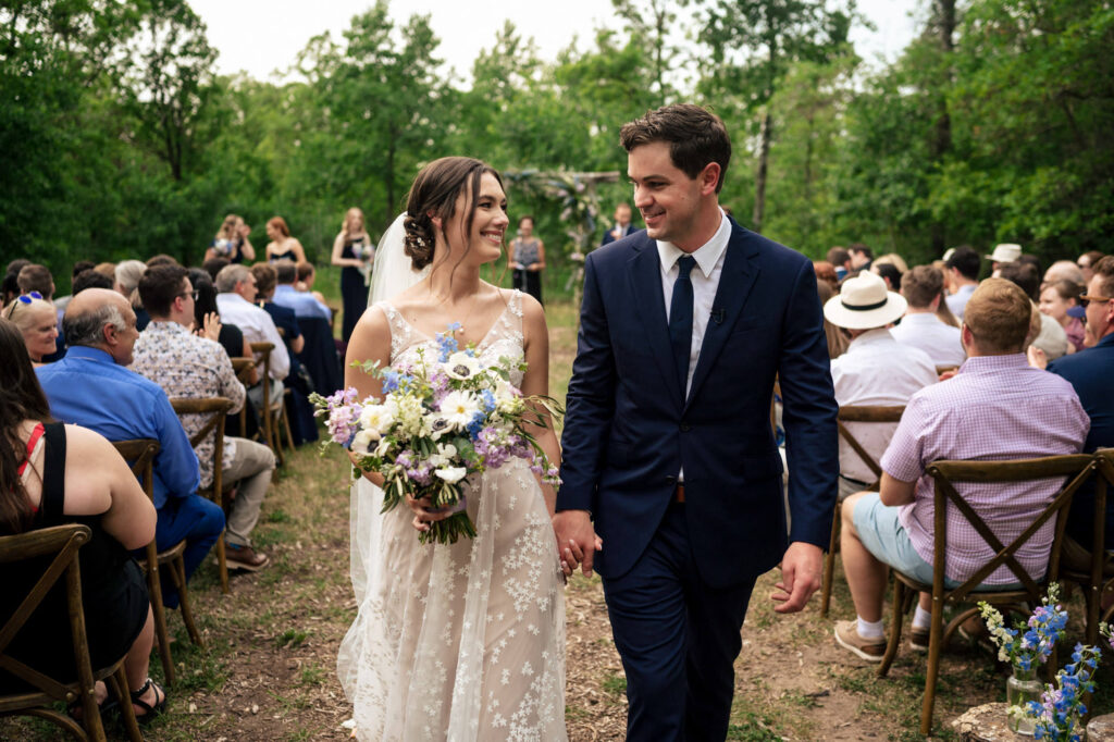 Bride and groom walk down outdoor aisle, holding hands and smiling.