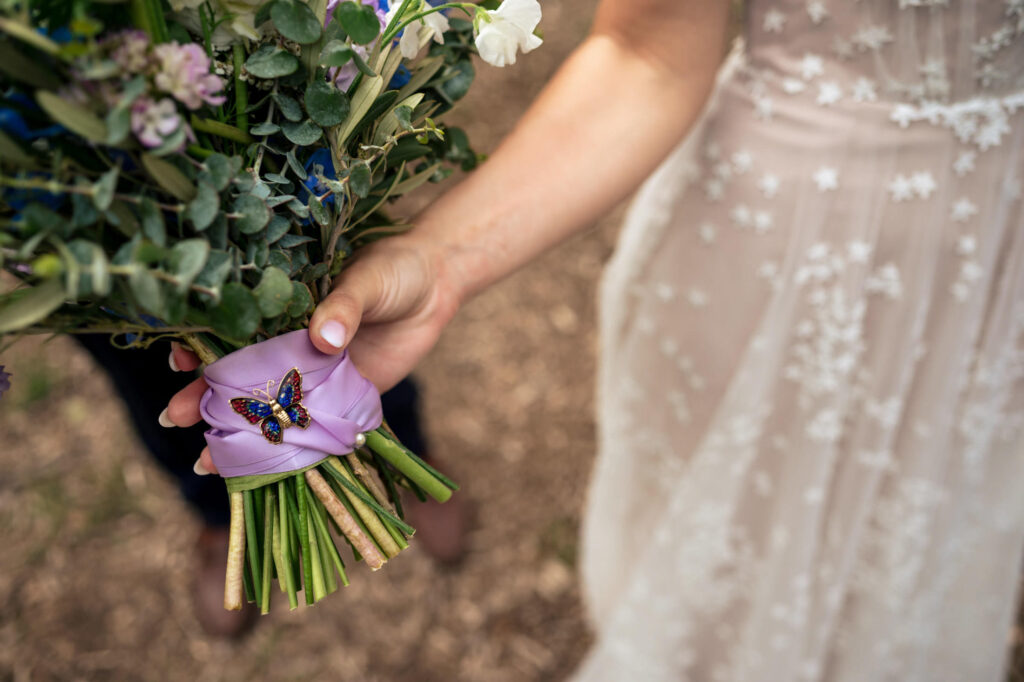 Bride holding bouquet wrapped in purple ribbon with a butterfly brooch, lace dress visible.