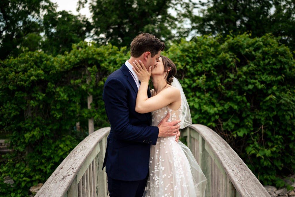 A couple kissing on a wooden bridge surrounded by greenery, with the woman in a white dress.