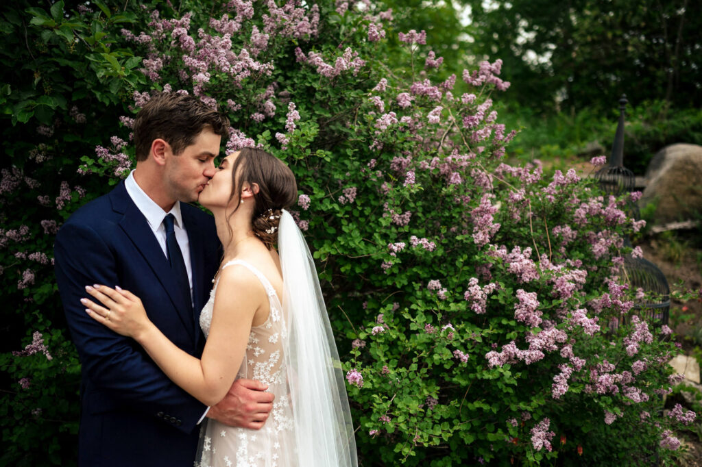 Bride and groom kissing in front of purple flowers and greenery.