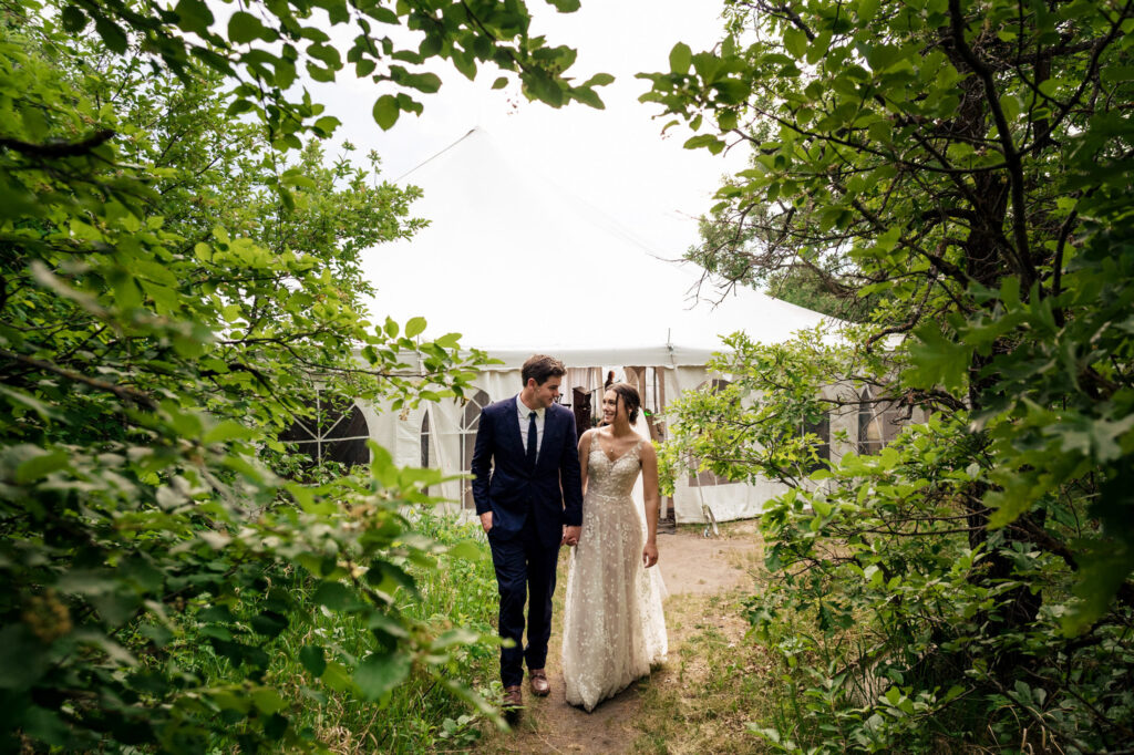 A couple in wedding attire walks through trees near a white tent.