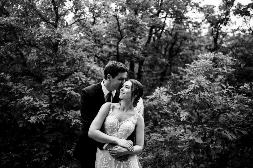 Bride and groom embrace in a forest, surrounded by trees.