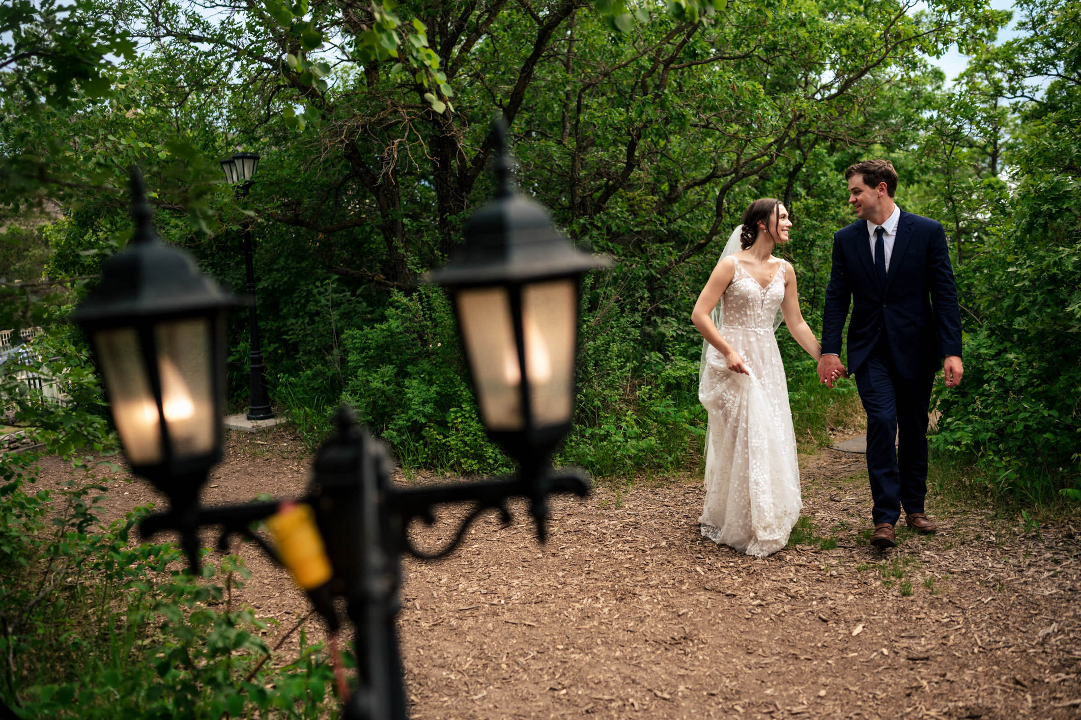 Wedding at Pineridge Hollow. A Bride and groom walking hand in hand on a wooded path, with lamp posts in the foreground.