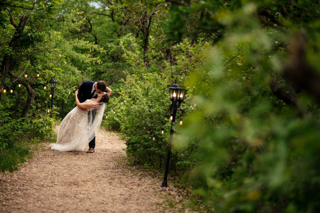 Bride and groom kissing on a wooded path with string lights and greenery.