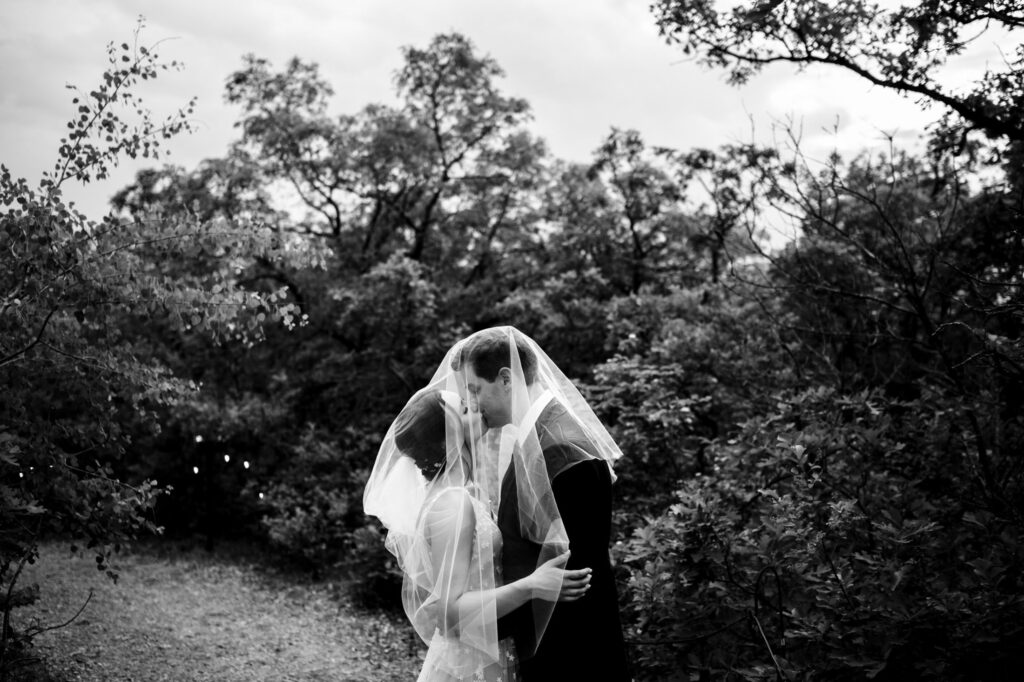 A couple kisses in a forest, both wearing wedding attire and veils.