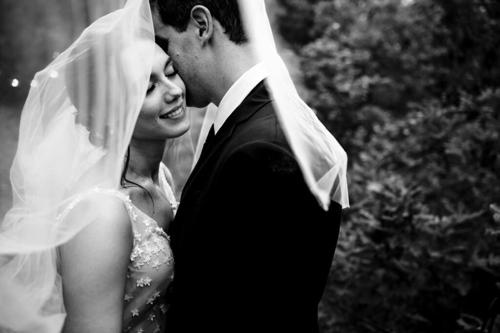 Bride and groom smiling under a veil in a forest setting, captured in black and white.