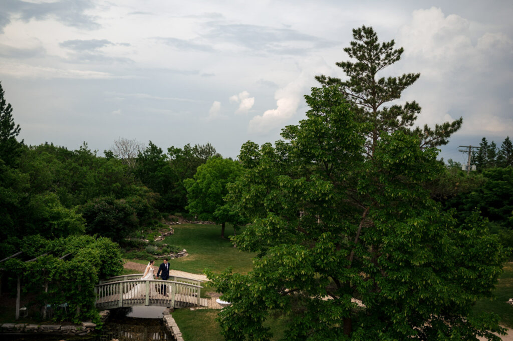 A couple walking on a small bridge in a lush, green park under a cloudy sky.