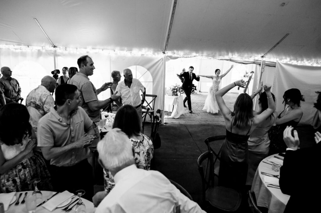 A couple joyfully enters a wedding reception tent as guests cheer and clap.