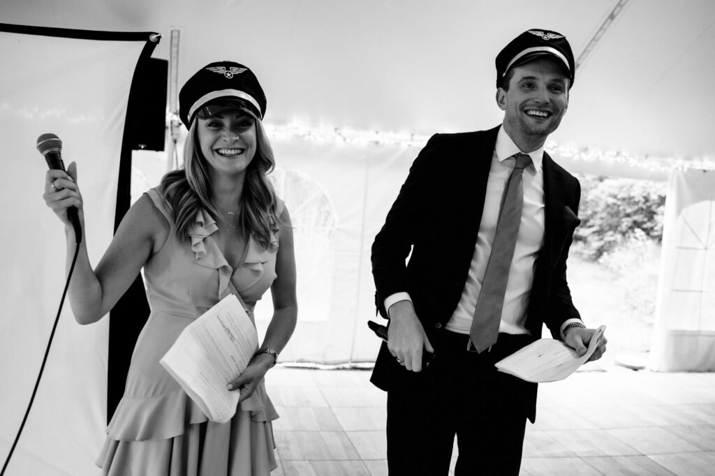 Two people in pilot caps, holding papers and a microphone, smiling under a tent.