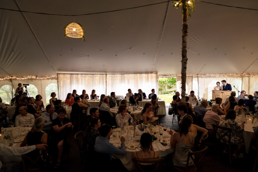 Wedding reception under a tent with guests seated and two people giving a speech.
