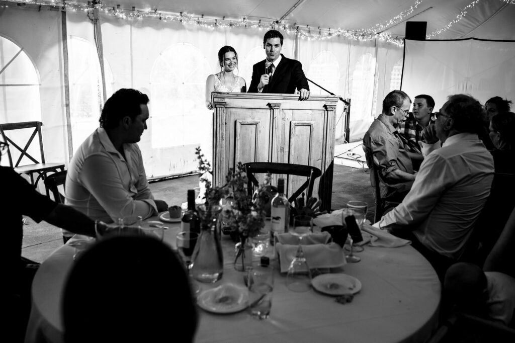 A couple speaks at a reception, guests seated around decorated tables.