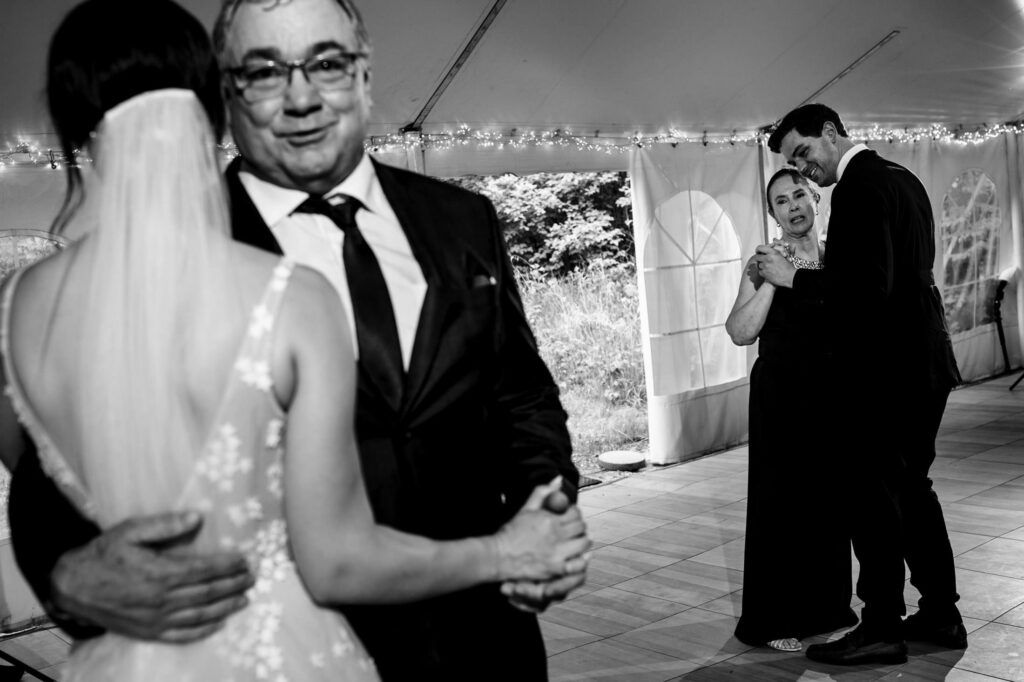 Two couples dance at a wedding reception under a tent with string lights.