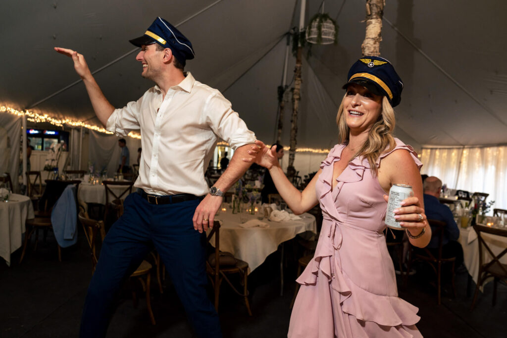 A man and woman in pilot hats dancing and laughing at a festive indoor event.