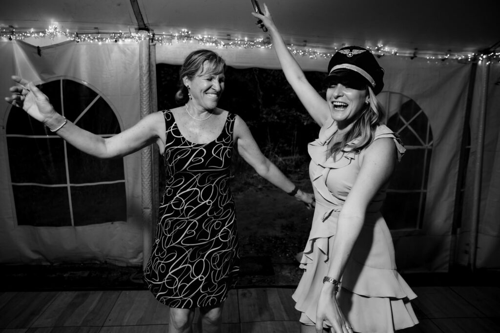 Two women dancing joyfully under string lights; one wears a dress, the other a pilot's hat.
