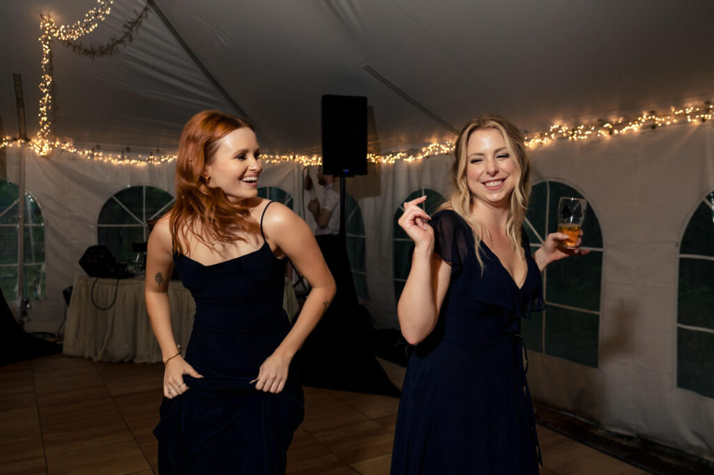 Two women in dark dresses dance and smile at an indoor party with string lights overhead.