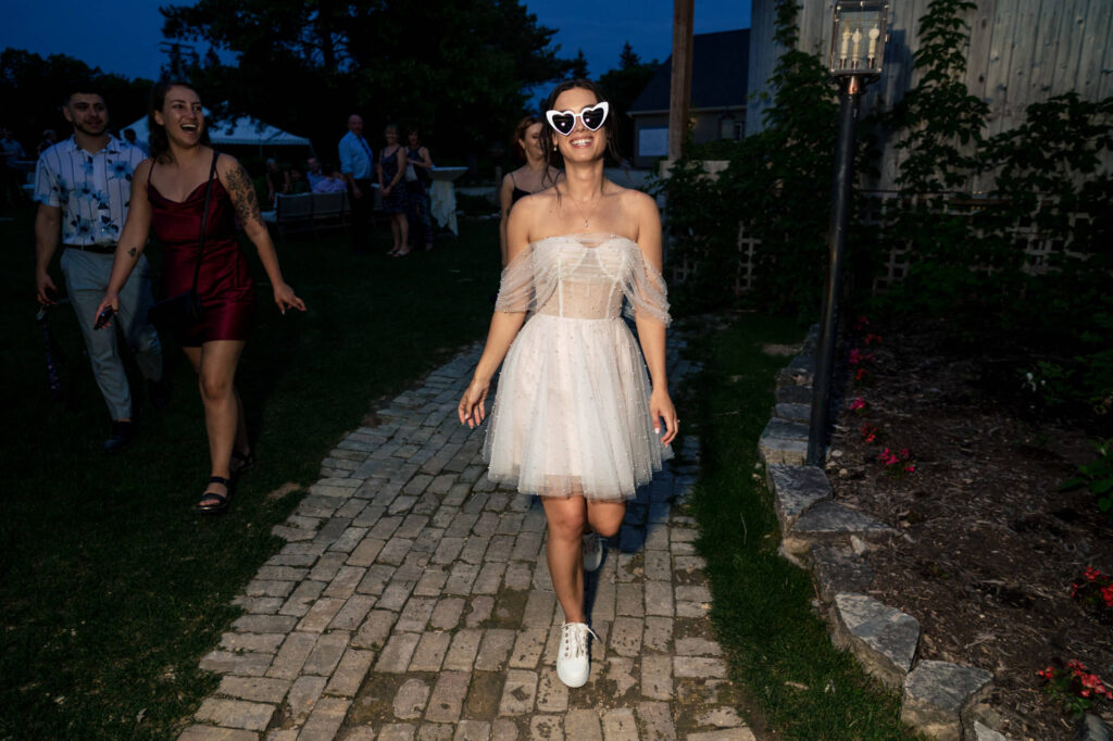 Woman in white dress and heart sunglasses walks on a brick path, followed by others at a gathering.