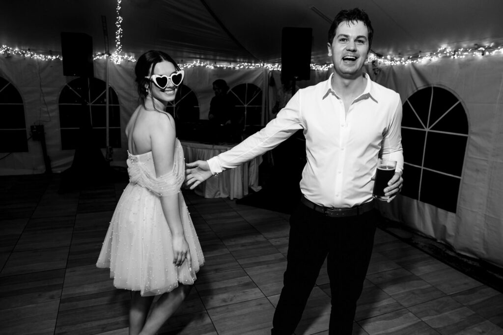 Couple dancing at a party, woman in heart glasses and man holding a cup, under string lights.