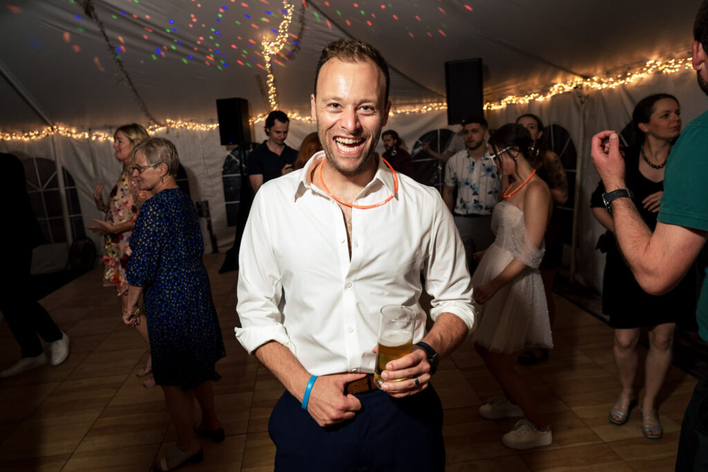Man smiling with a drink on a dance floor at a party; festive lights in the background.