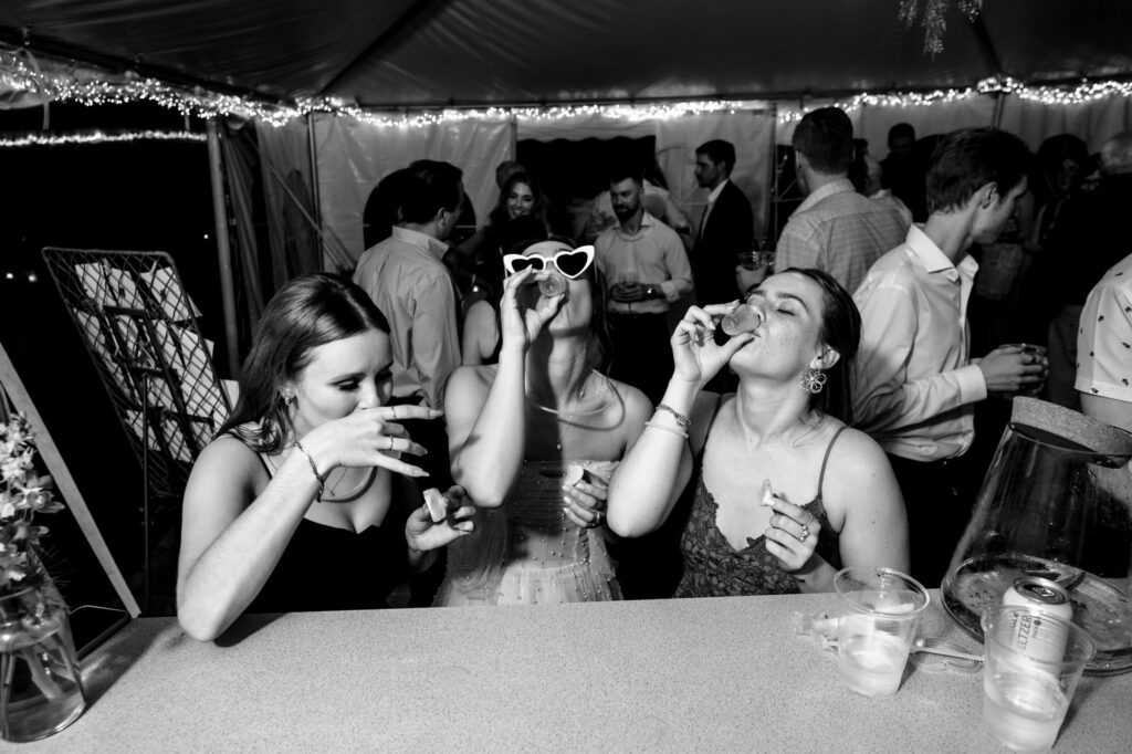 Three women at a party taking shots, one wearing heart-shaped sunglasses, surrounded by people.