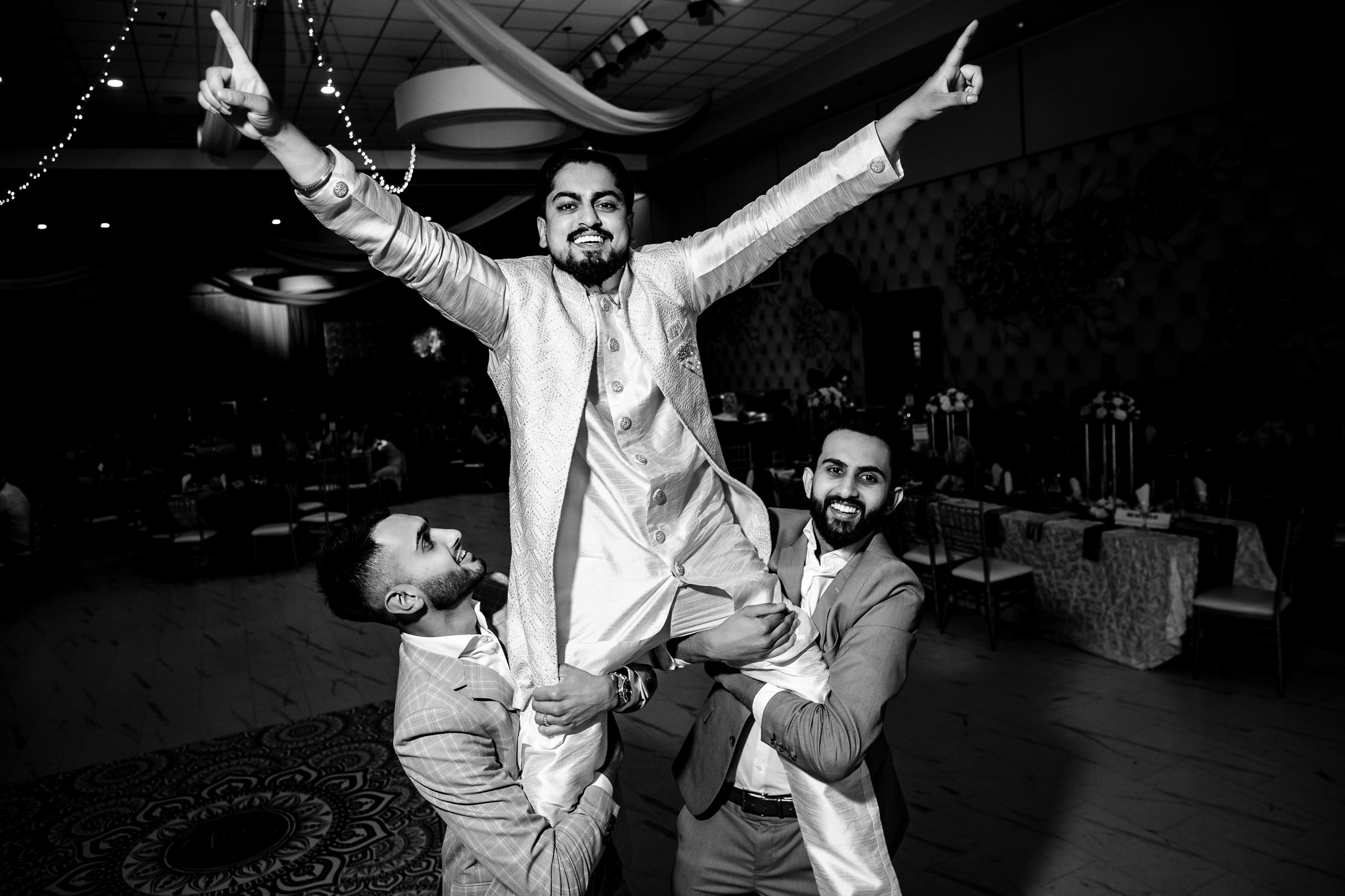 Three men in suits hold up a joyful man with raised arms in a decorated hall.