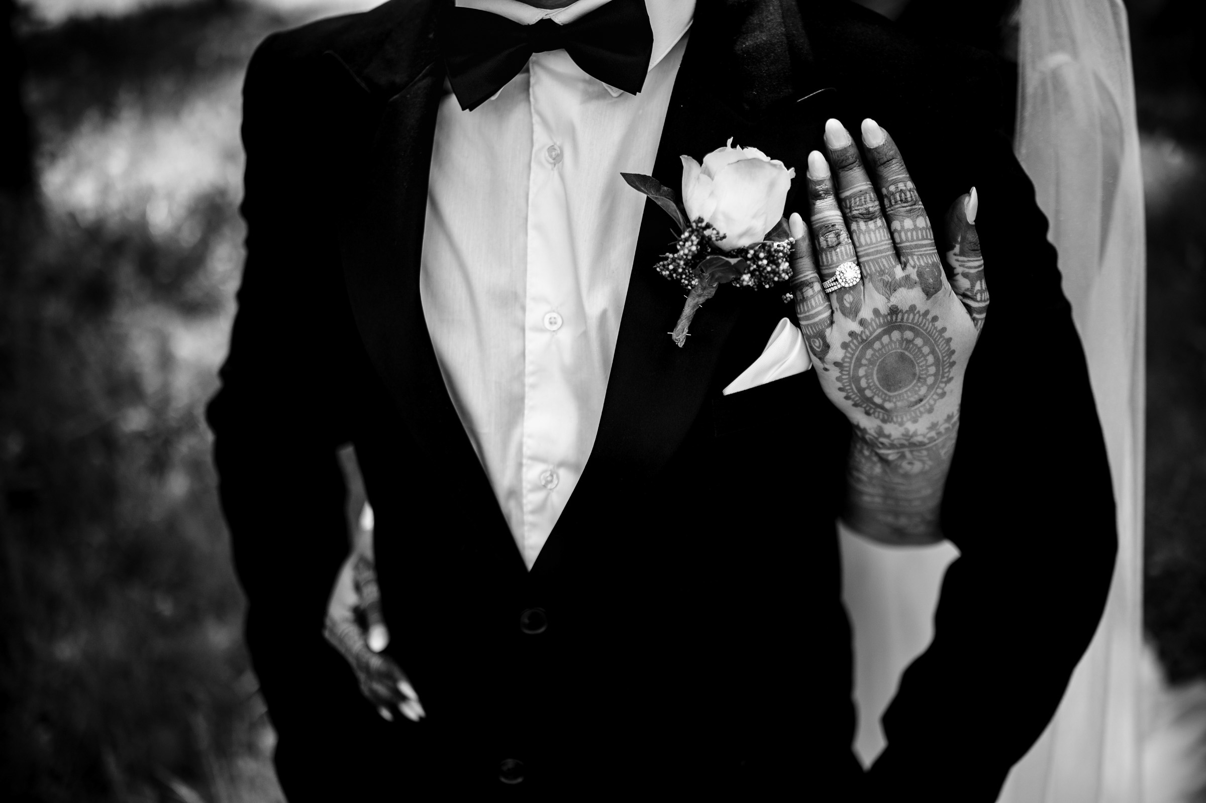 Bride in veil embraces groom in suit; henna-decorated hand with rings rests on his chest.