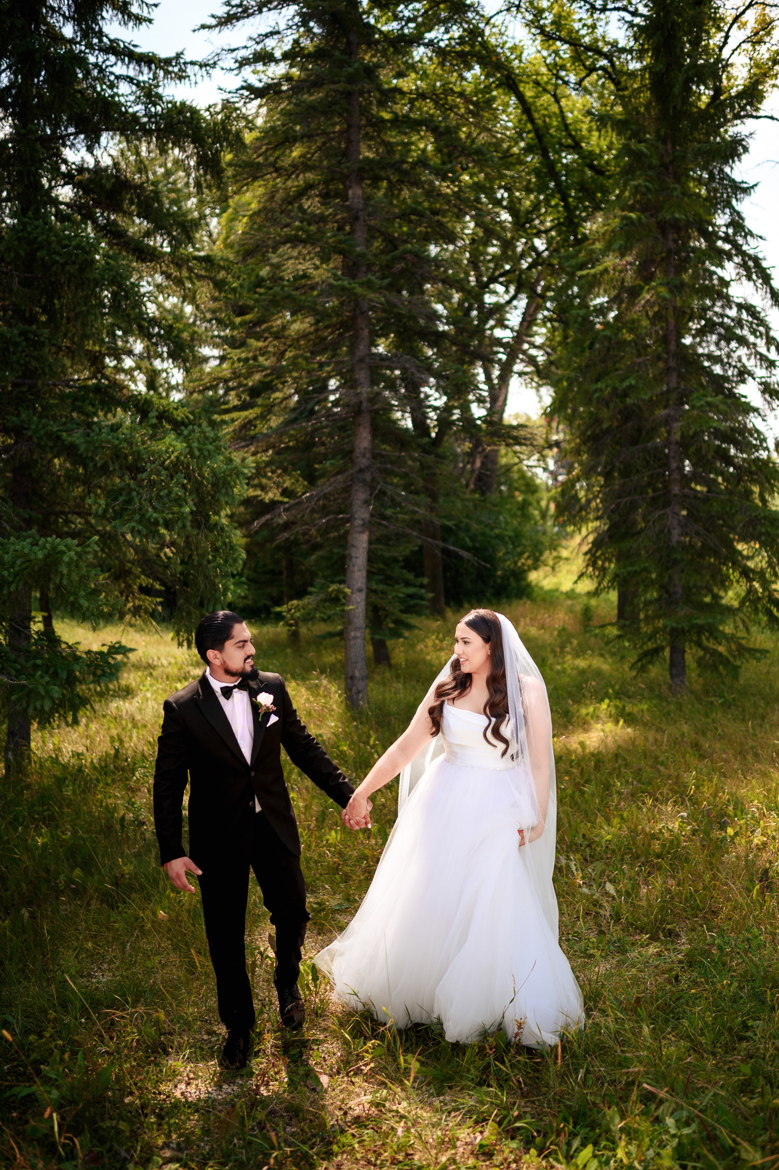 A bride and groom hold hands, walking in a forested area.